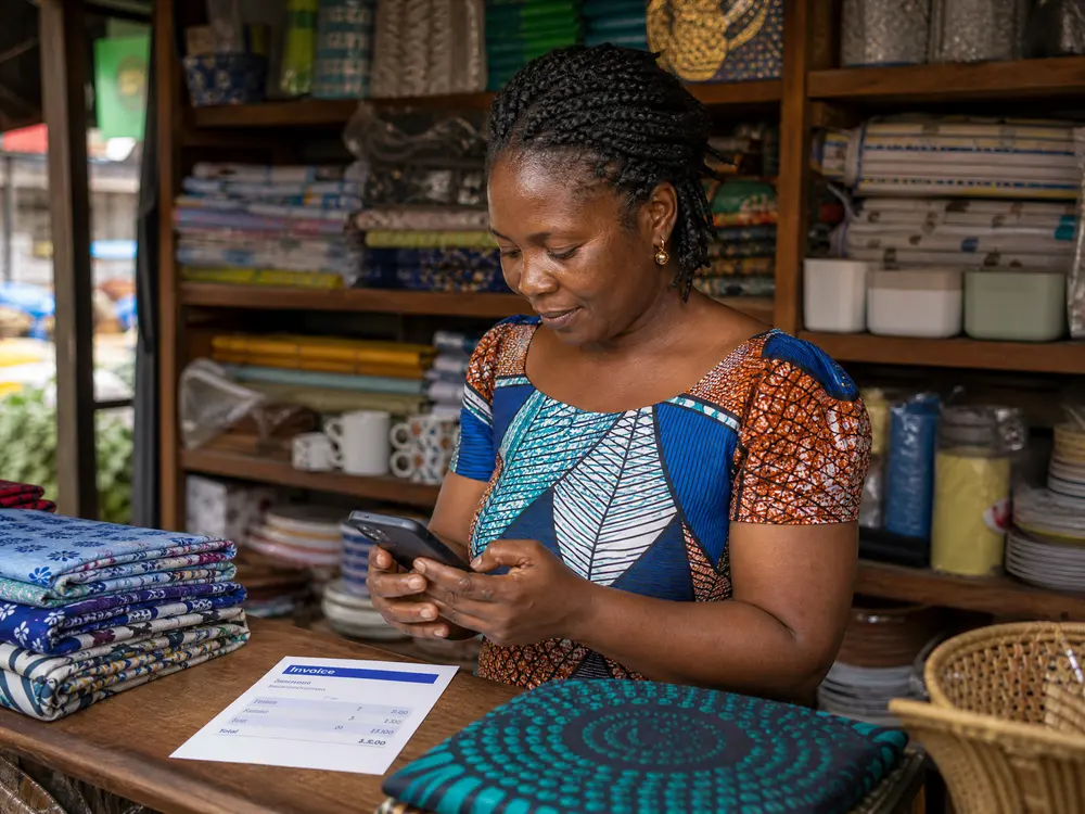 A Ghanaian shop owner in her fabric and homeware store checking an invoice on her phone at the counter.